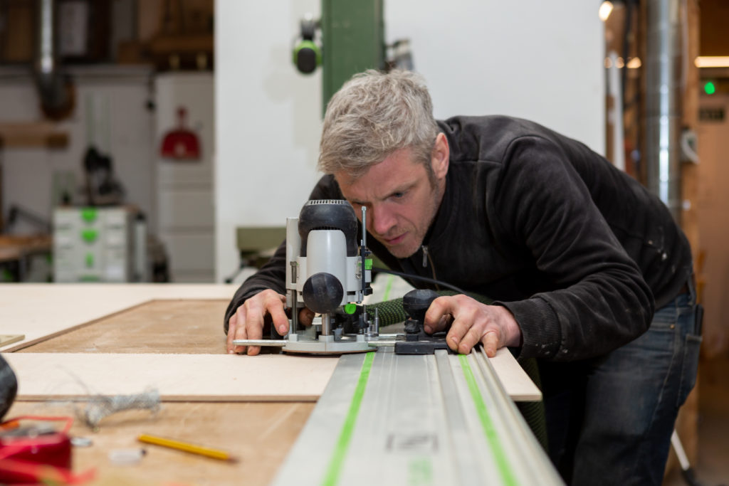 A man is using a router on a large sheet of wood clamped to a work bench