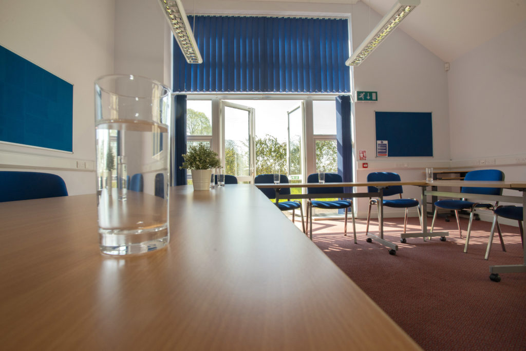 Williton enterprise centre conference space. Desks are arranged around the outside of the room with a space on the inside. There are class doors leading to an outdoor space. There are blue pin boards mounted on two walls.