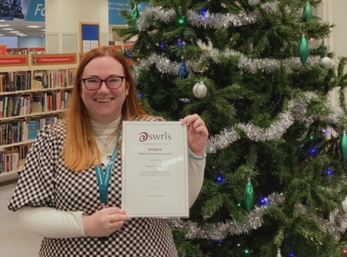 Christina Evans, Library Manager and Development Officer, accepting the SWIRLS CIO Initiative Award 2023 stood in front of a Christmas tree in a library.