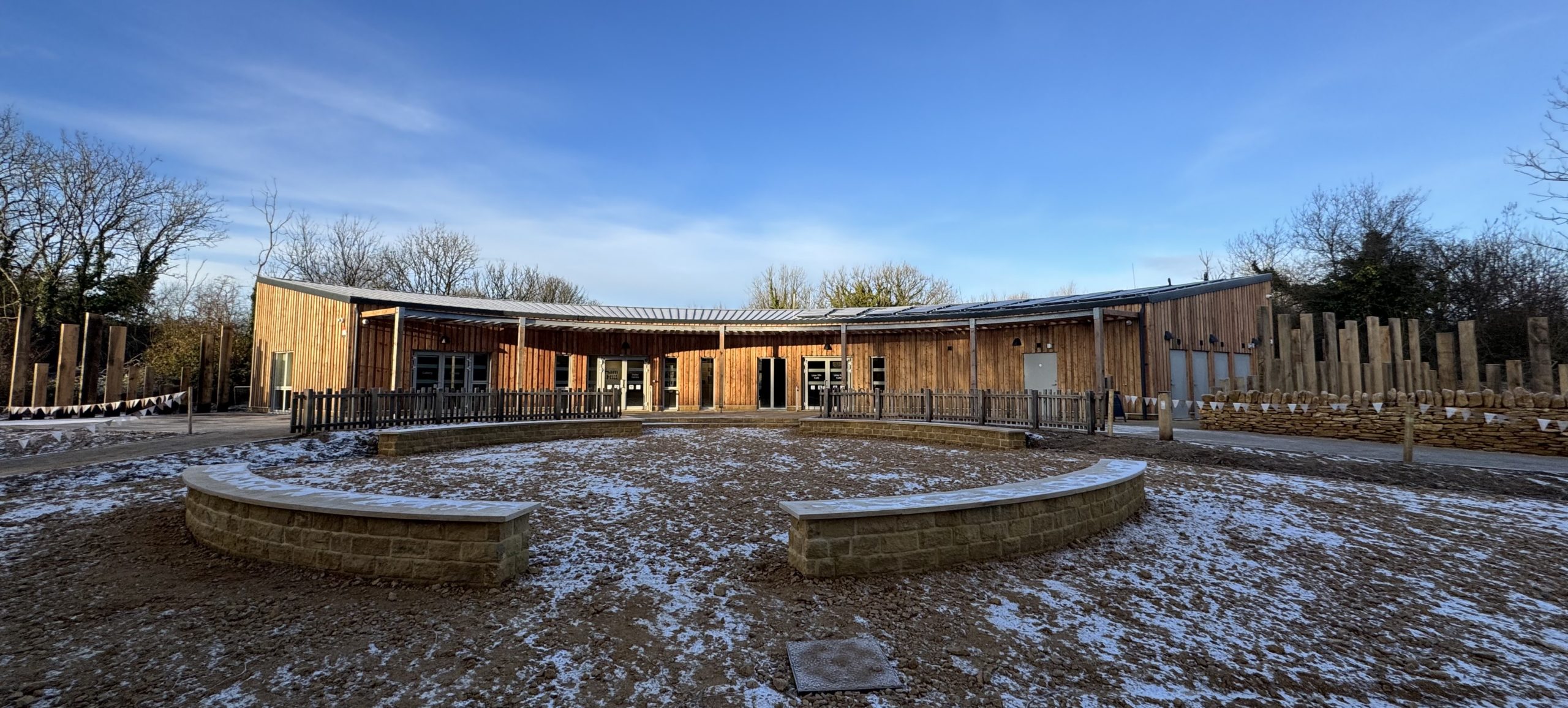 A wooden, single‑storey visitor centre curves around a circular outdoor space dusted with light snow, with trees and a bright blue sky in the background