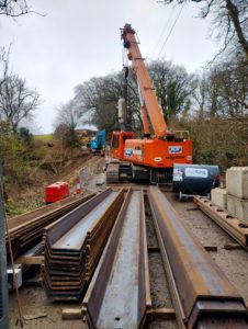 A crane hoists steel beams onto a track at a construction site, with a road visible in the background.