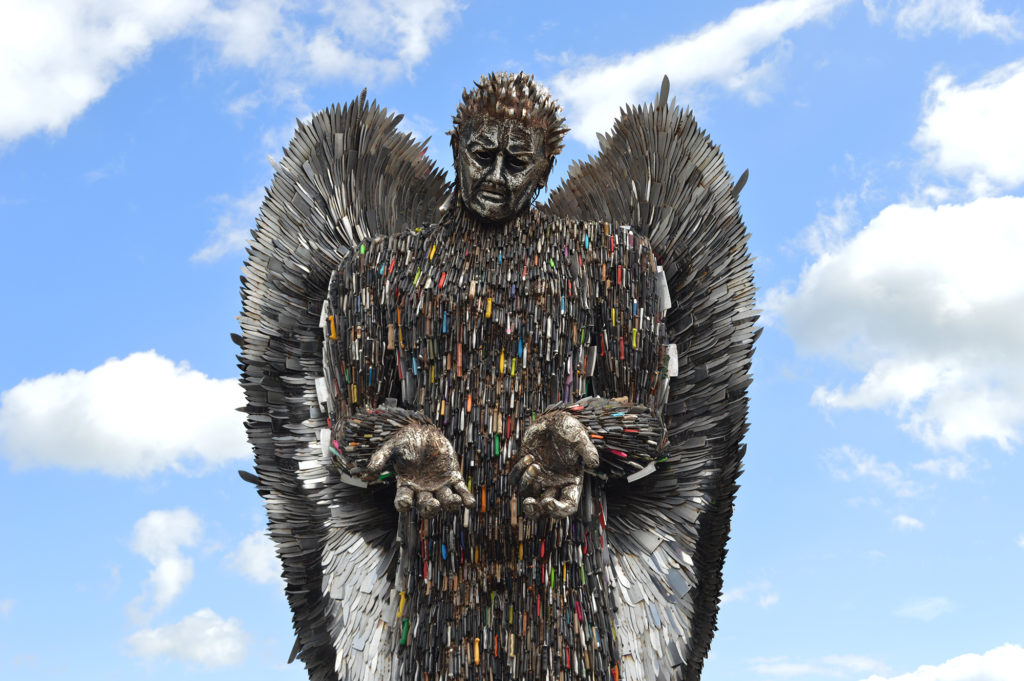 Giant statue of an angel made out of knives stood in front of a blue, cloudy sky.