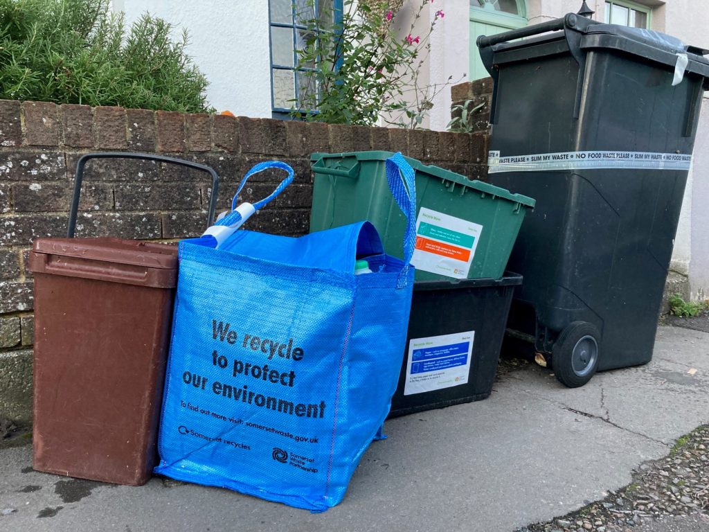 A brown caddy, blue bag, green and black recycling boxes and a black wheelie bin filled with waste next to the front wall of a house on the pavement.