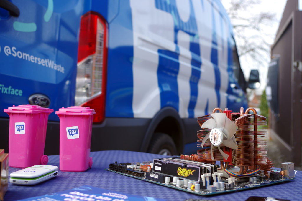 2 pink, Fixy-branded miniature wheelie bins with a mobile and PC motherboard on a table, with Fixy van in the background.