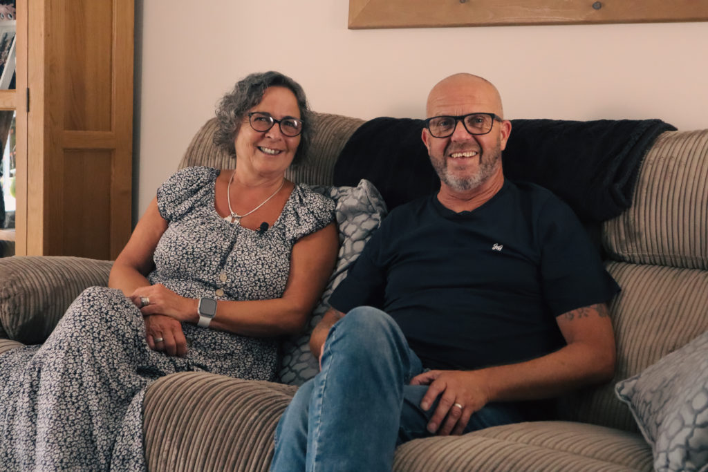 Foster parents Amanda and Steve, smiling whilst sat on their sofa.