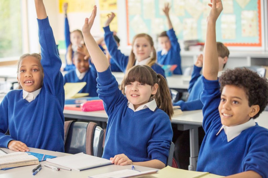 Students in blue uniforms raise their hands in a classroom, with notebooks and supplies on desks.