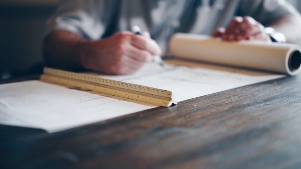 A planner using a pen and ruler whilst looking at a scroll of development plans.
