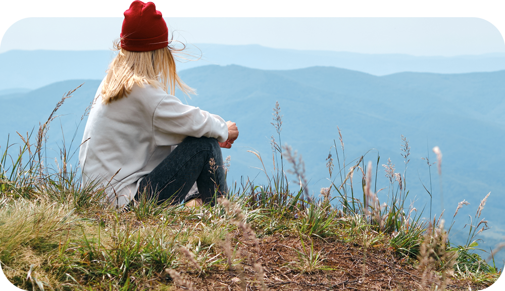 A woman in a red beanie and white sweater sits cross-legged on a grassy hill, gazing at distant blue mountains.