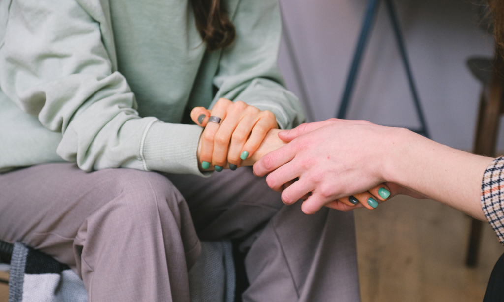 Two people sitting close, holding hands with green nail polish; one wears a light green sweater and grey trousers.