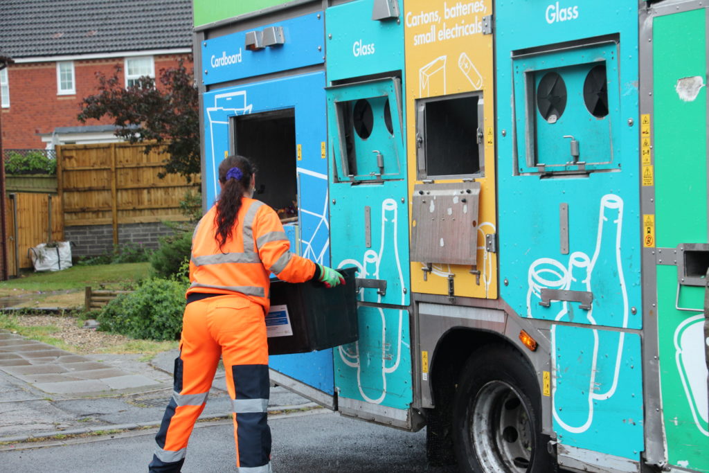 Recycling crew member in high-visibility gear loads sorted materials into a colourful recycling truck on a residential street.