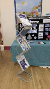 A collapsible zig0-zag leaflet stand is holding 6 different bits of information in front of a table display stand at an event.