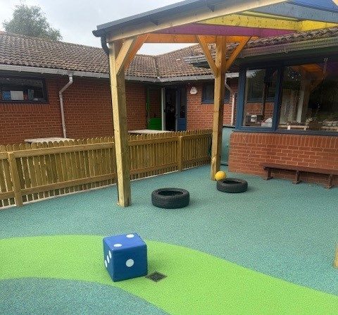 A play area with a wooden pergola with a colourful roof. There is a green pattern on the ground, a giant dice, tires and a ball.