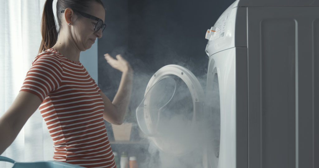 Upset woman staring at her broken washing machine, the room is full of smoke.