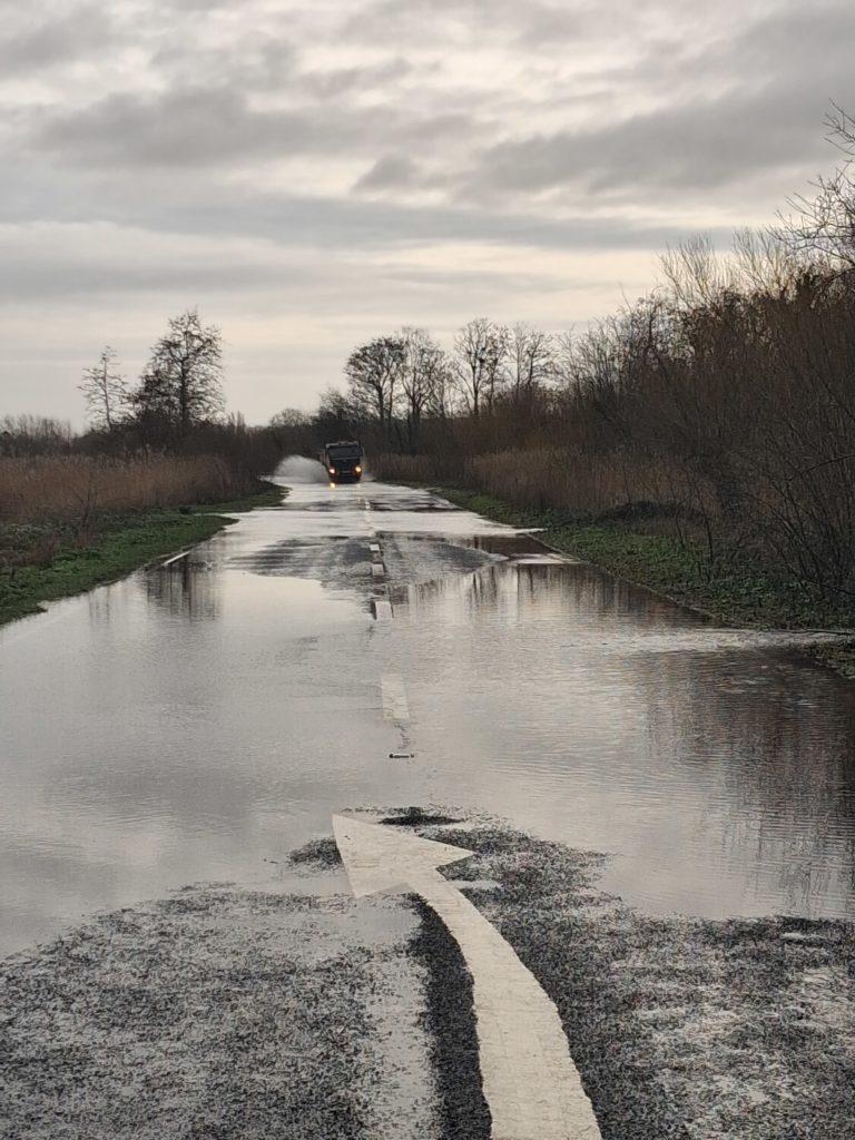 Flooded section of the A361 with standing water covering the road, a white arrow marking visible, and a vehicle stopped in the distance.