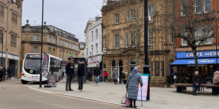 People walking and sitting along Yeovil High Street, with shops, cafés, historic buildings, and a bus stopped at the roadside.
