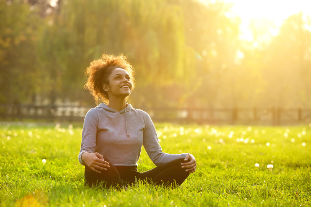 Happy young woman sitting in yoga position in a sunlit park, meditating on green grass surrounded by trees.