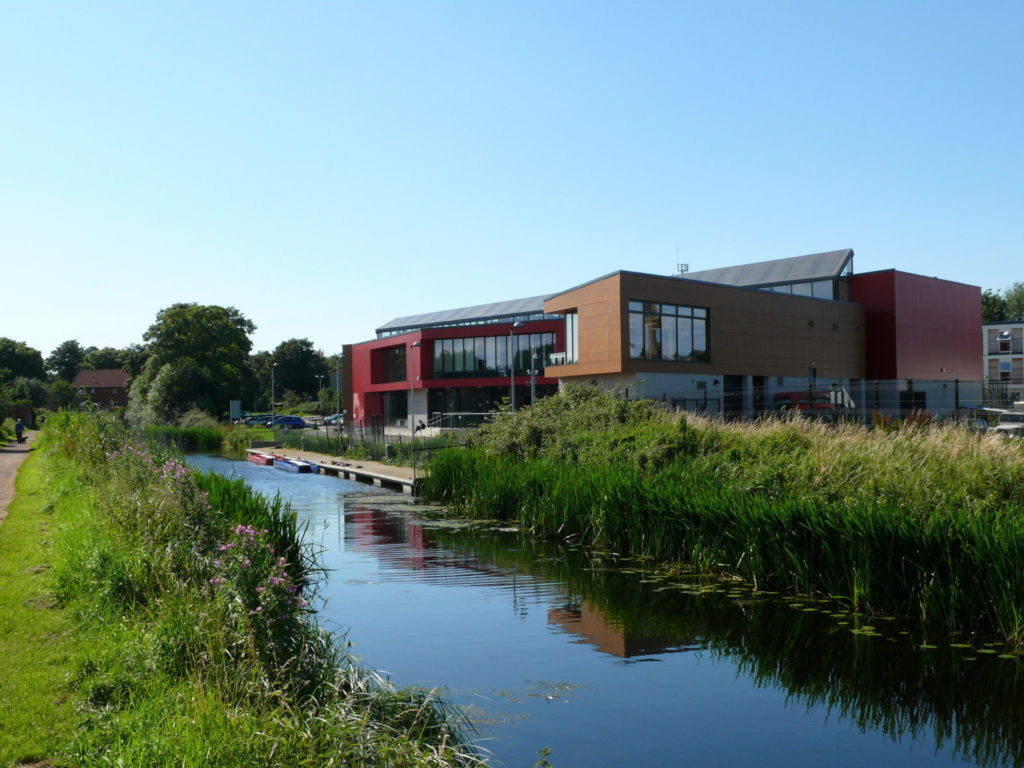 Modern waterside building beside the Bridgwater and Taunton Canal, with reeds and calm water reflecting the structure under a clear sky.