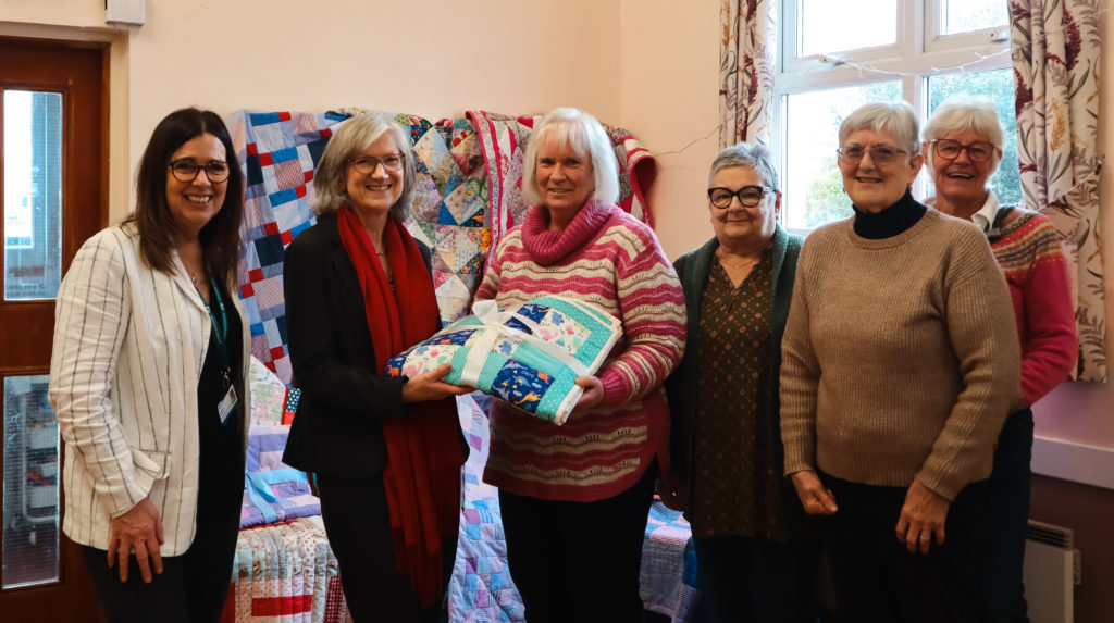 Six women standing in a row with blankets behind them. Two women in the middle jointly holding a folder up blanket.