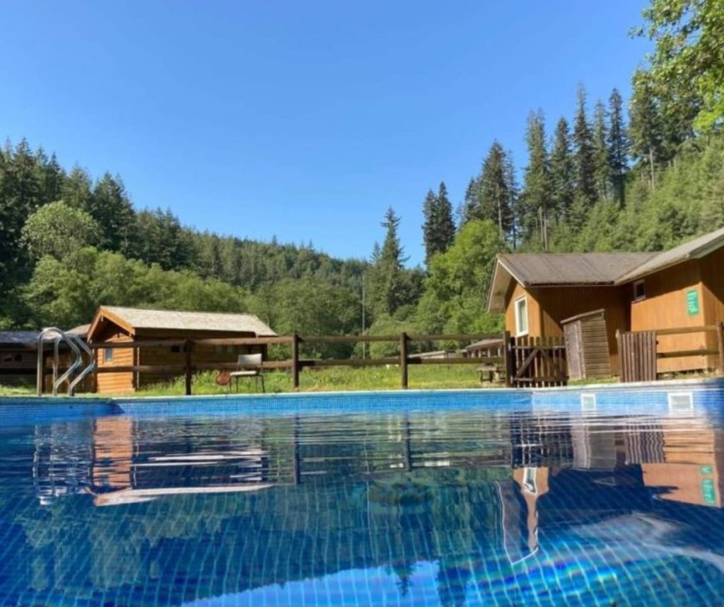 Outdoor swimming pool beside wooden cabins, with forested hills and clear blue sky in the background.