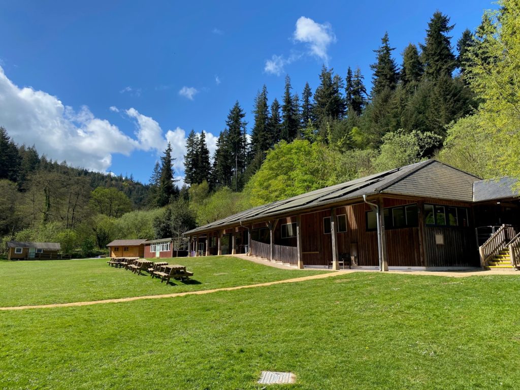 Wooden lodge building with picnic benches on a grassy field, surrounded by tall trees and hills under a blue sky.