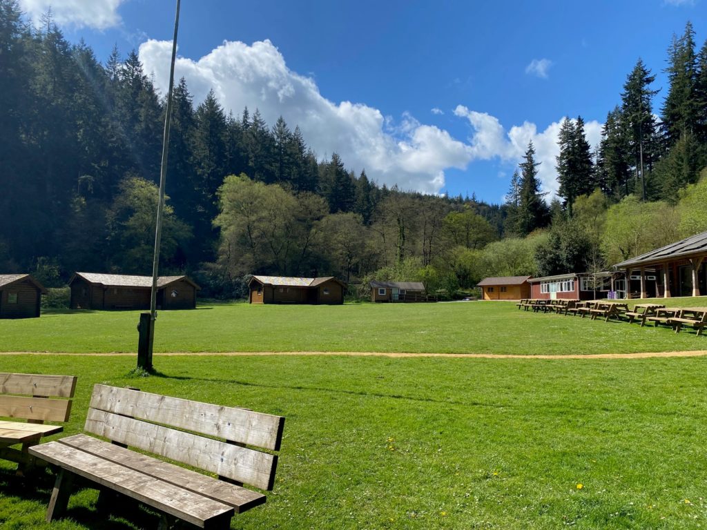 Open grassy campsite with wooden cabins, picnic benches, and forested hills under a partly cloudy sky.