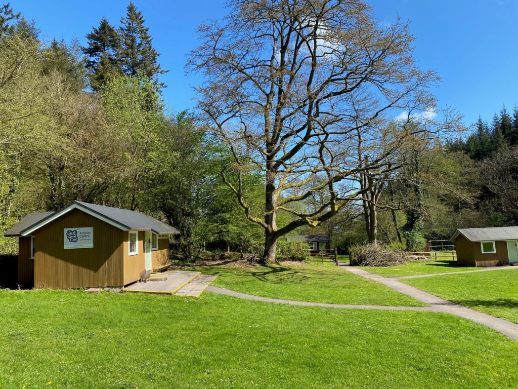 Small wooden building with a “Great Wood Camp Activity Centre” sign, set on a grassy area with a large tree and surrounding woodland.