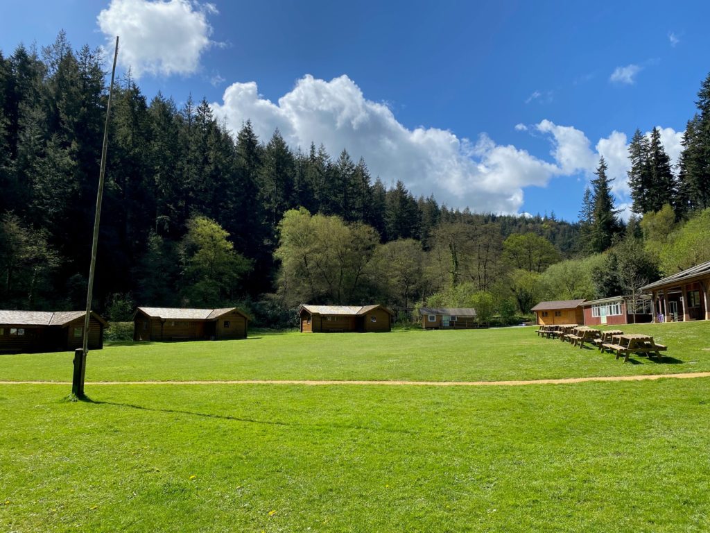 Grassy campsite with several wooden cabins and picnic benches, backed by dense forest and a partly cloudy blue sky.