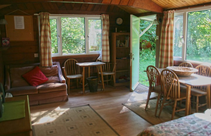 Interior of a wooden cabin with a table, chairs, sofa, and open door leading to a green outdoor area.