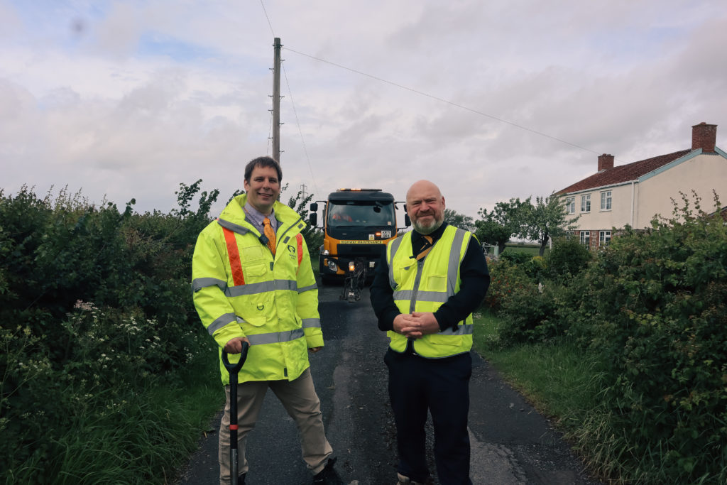 Cllr Wilkinson (left) and Cllr Revans stand on A30 Hendford Hill with highways vehicle behind during repair works.