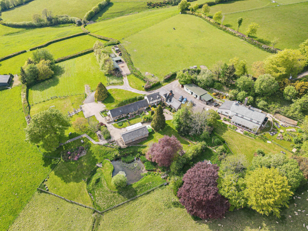 Aerial view of a rural property with houses, gardens, and surrounding green fields divided by hedges.