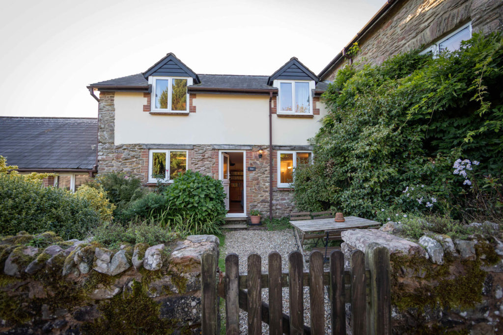 Stone cottage with an open front door, surrounded by plants and a small garden with a wooden table and fence.