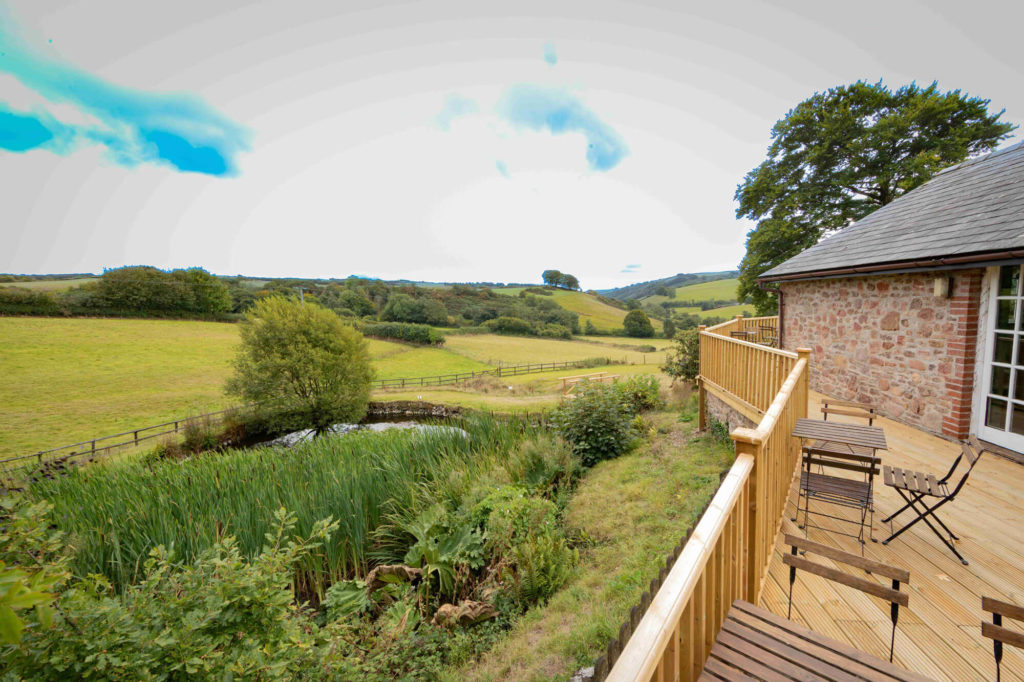 Balcony with outdoor table and chairs overlooking a pond, surrounded by greenery and open countryside.