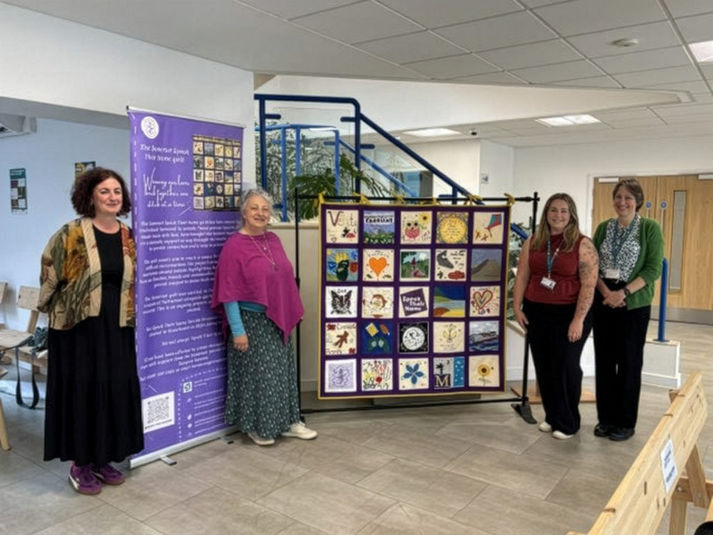 Group stands beside the Speak Their Name Quilt and banner in indoor space, highlighting community memorial artwork project.
