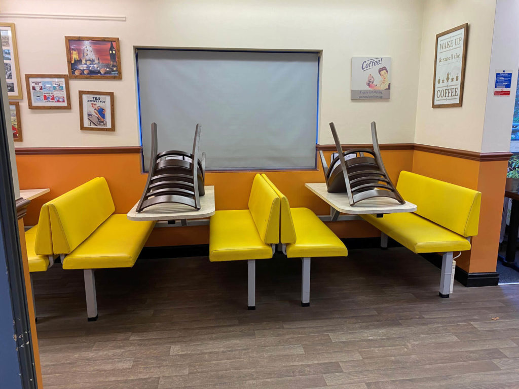 Cafe seating area with yellow booths and chairs stacked upside down on tables against a wall with framed posters.