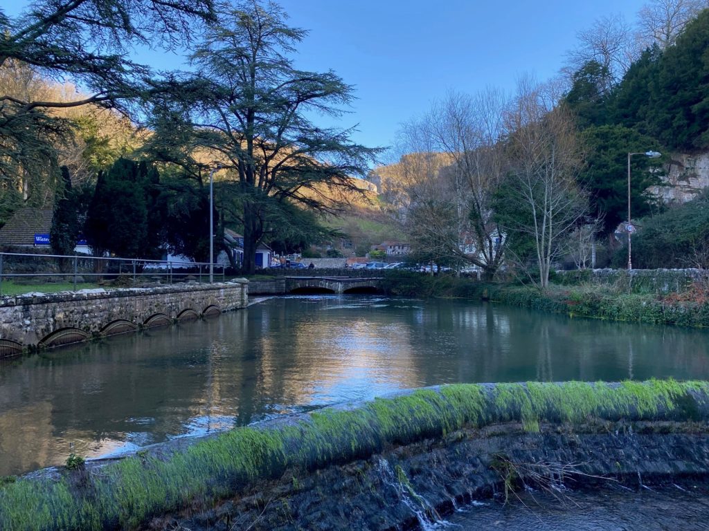 River with a small stone bridge and trees along the banks, with buildings and hills in the background.