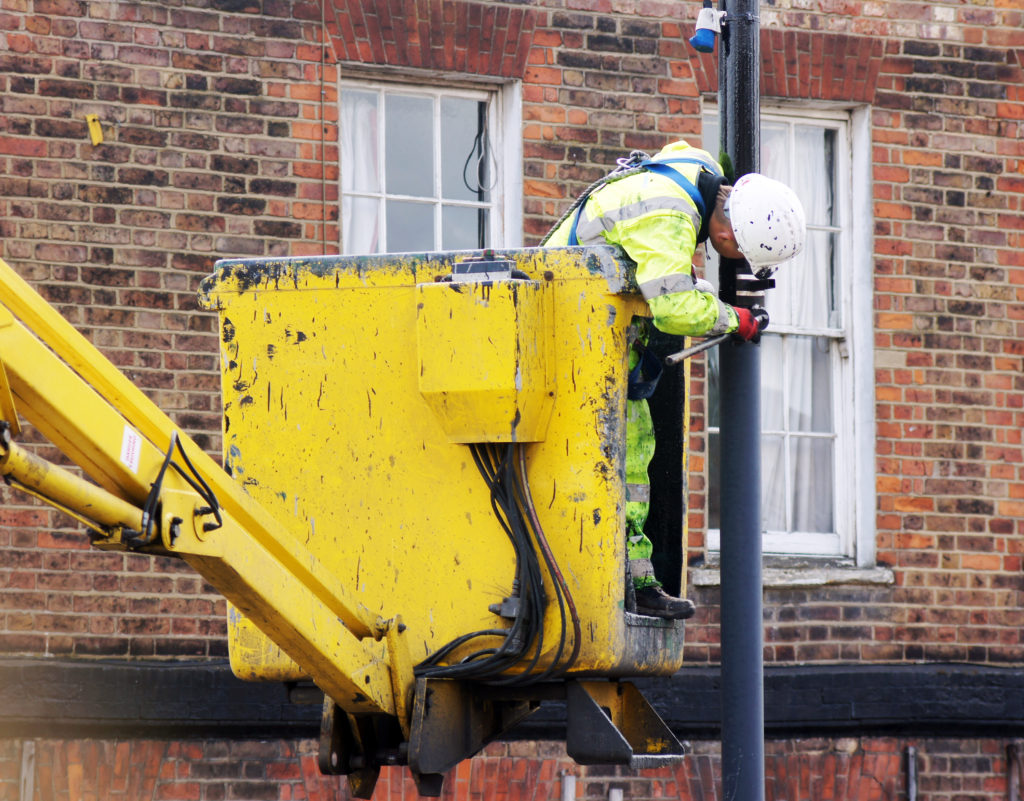 Worker in high-visibility clothing uses a raised platform to maintain or install equipment on a street pole outside a brick building.