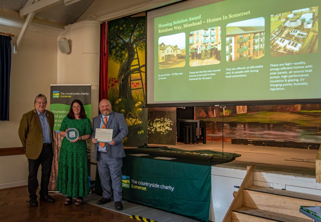 Cllr Smith-Roberts (centre) and Cllr Revans (right) receive CPRE Somerset award on stage with housing project presentation in background.