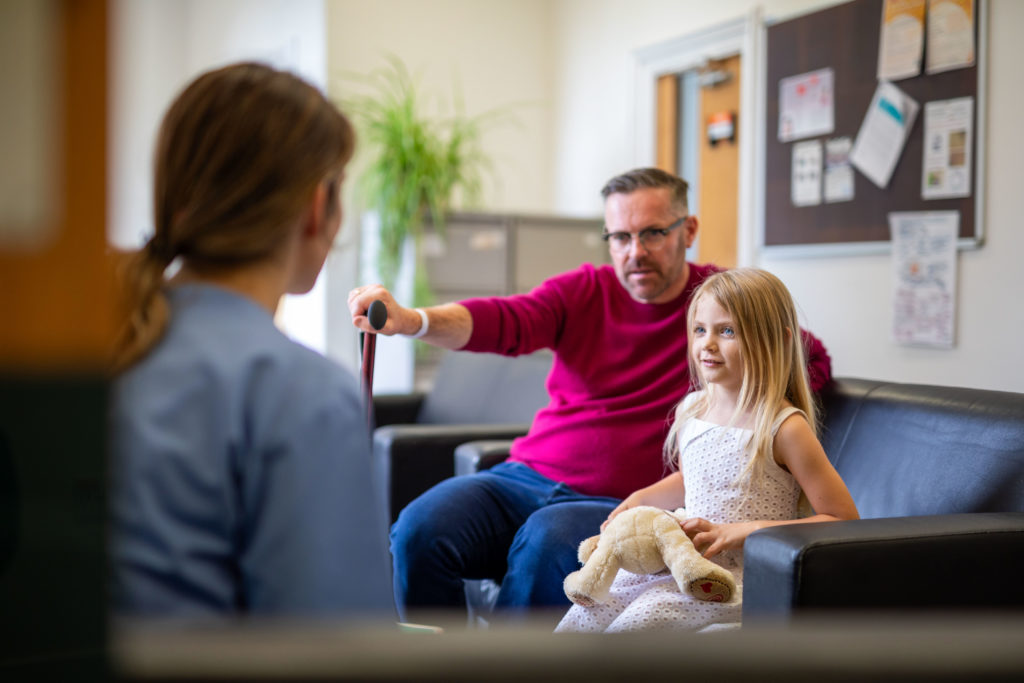 Father and young girl with soft toy speaking to support worker in waiting room during family support or social care meeting.