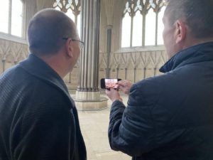 Two men stand in the ornate Chapterhouse at Wells Cathedral. One is showing the other an image on a smart phone.