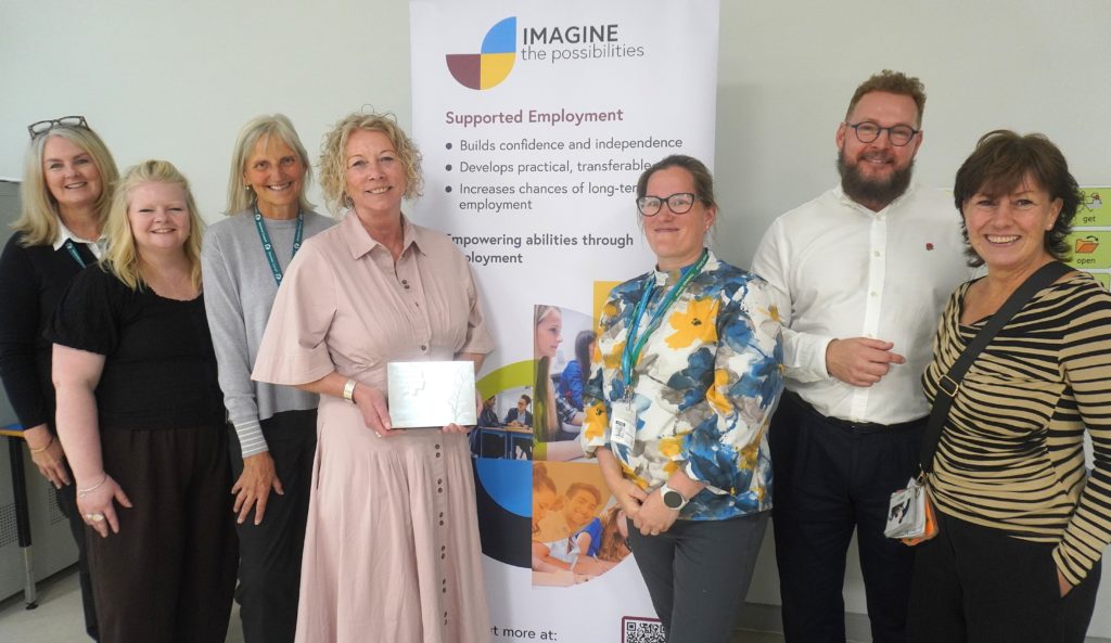 Imagine the Possibilities team holding award plaque at Fiveways School, standing in front of supported employment banner.