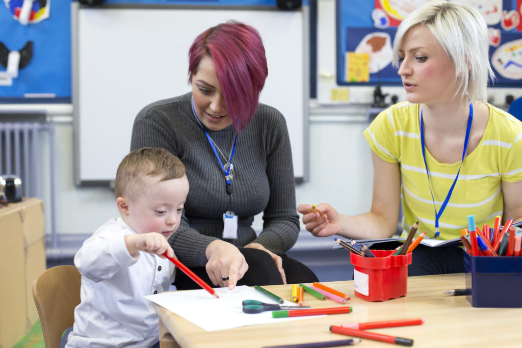 Adult and young child drawing together at a table with support from two staff in a classroom setting.