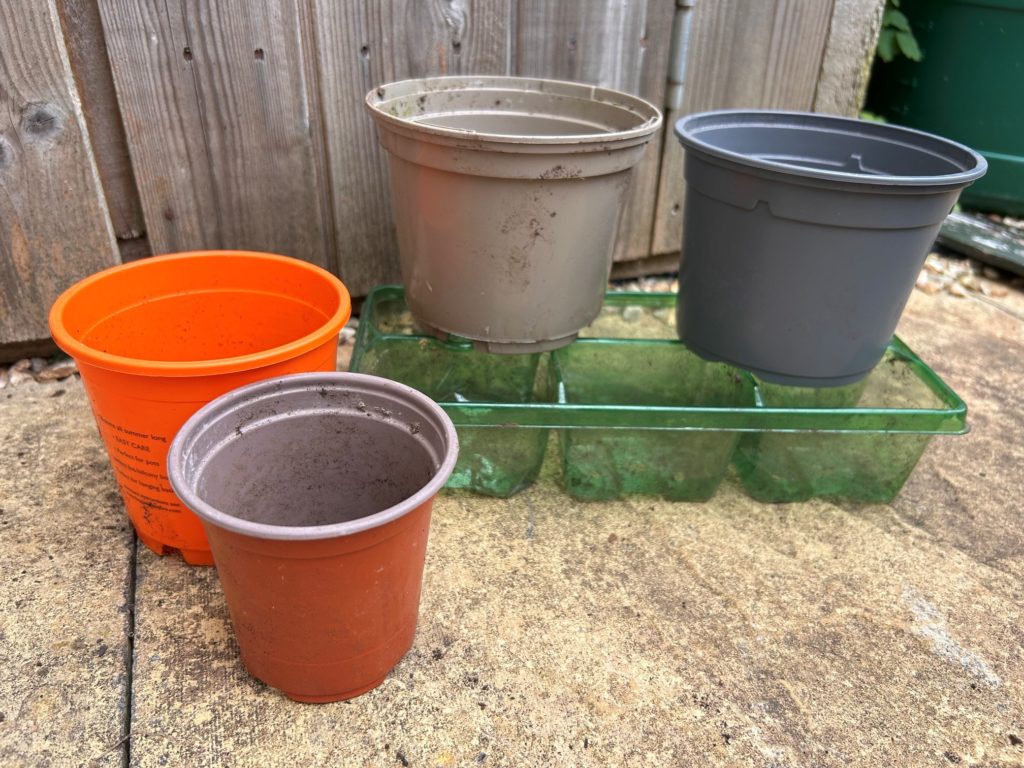 Selection of used plastic plant pots in various colours and sizes, including orange, grey and brown, placed outdoors on a patio.