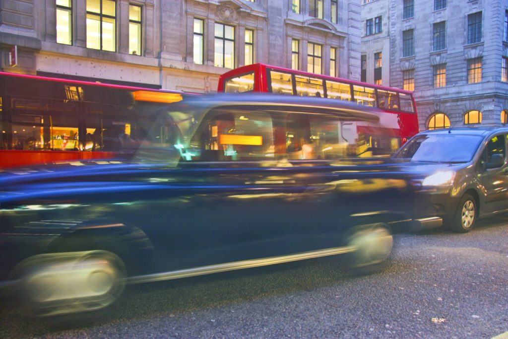 Motion-blurred hackney carriage travelling through a city street, with buses and illuminated buildings in the background.