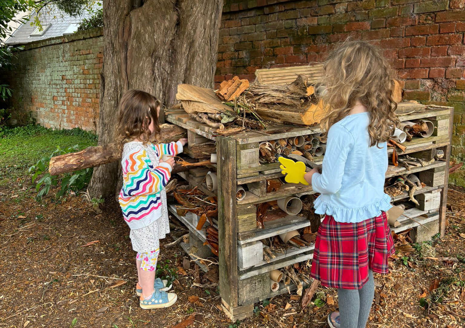 Two young children are adding sticks to a large pallet bug hotel in a churchyard next to a tree.