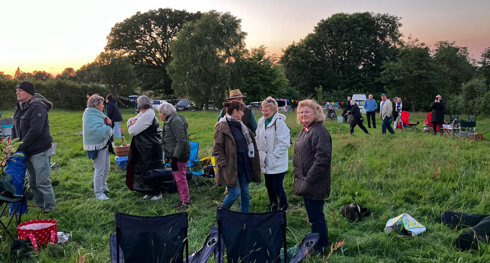 Several people are gathered in small groups in a meadow at sunset. There are camping chairs and tables scattered around.
