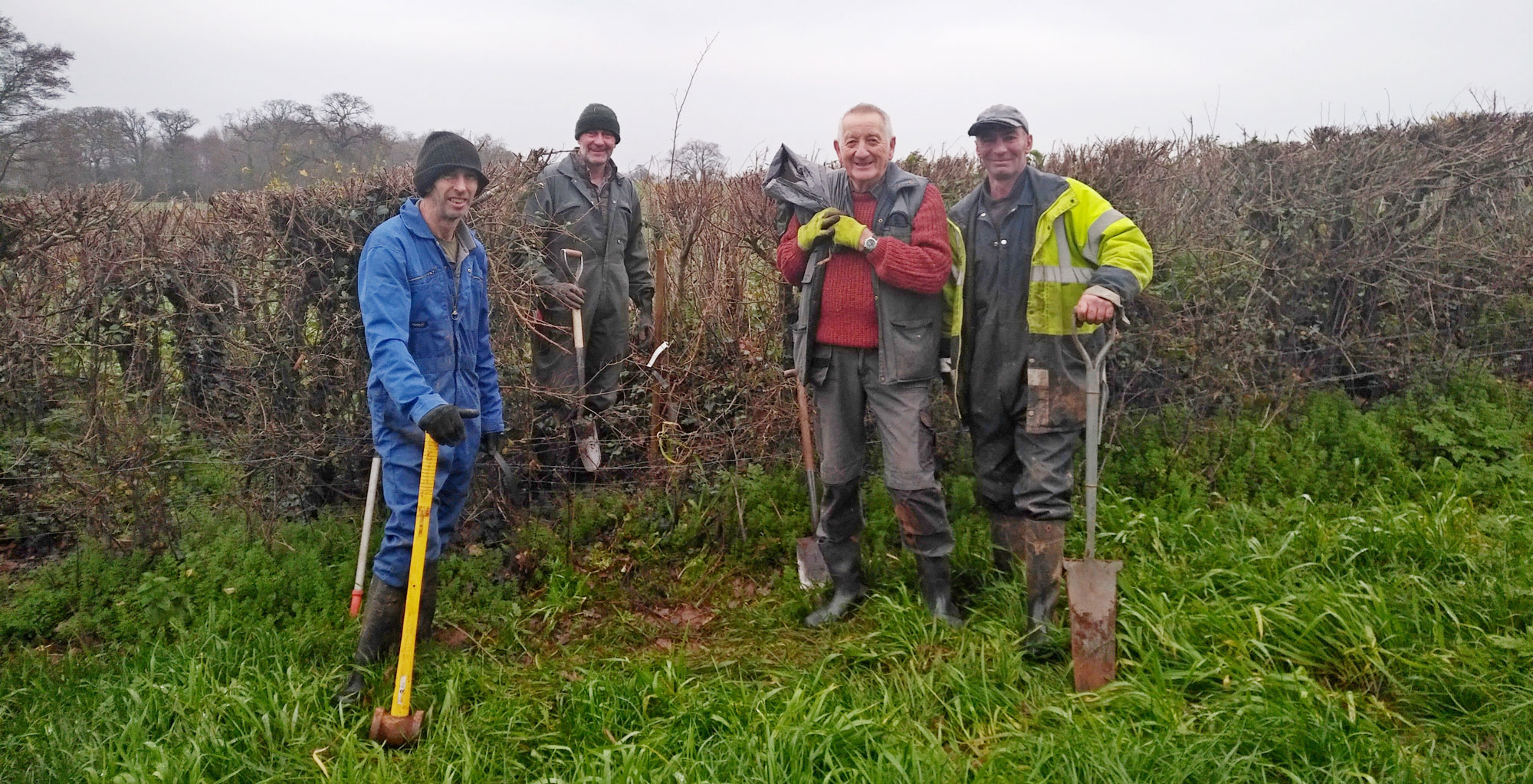 4 men in overalls are standing next to a hedge, they are all carrying hedge planting tools