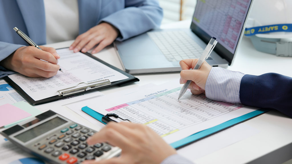 Finance professionals reviewing accounts, audit reports and budget figures around a desk with a laptop and calculator.