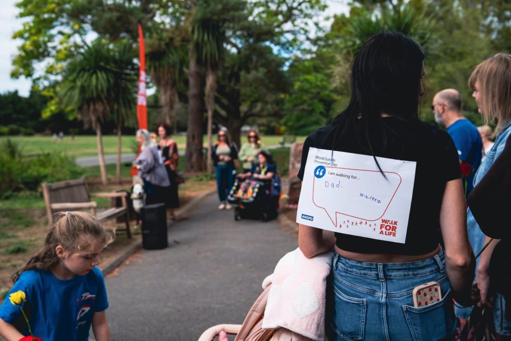 Participant at a Walk for Life event wears a tribute message for a loved one while people gather in a park.