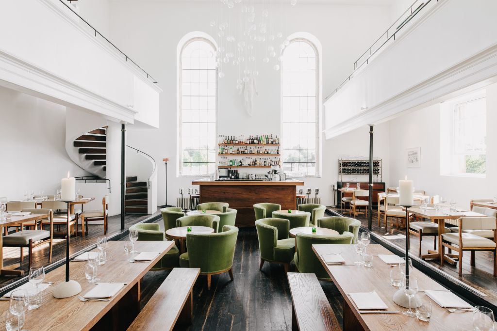 Light-filled restaurant and bar area within a converted chapel, featuring high arched windows, a central wooden bar, green armchairs, dining tables, and a sweeping staircase.
