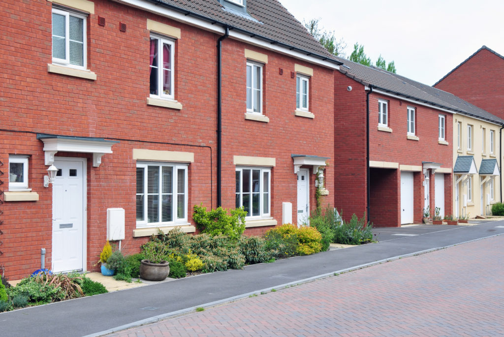 Modern red-brick terraced homes with front gardens, driveways and garages on a newly built residential street.